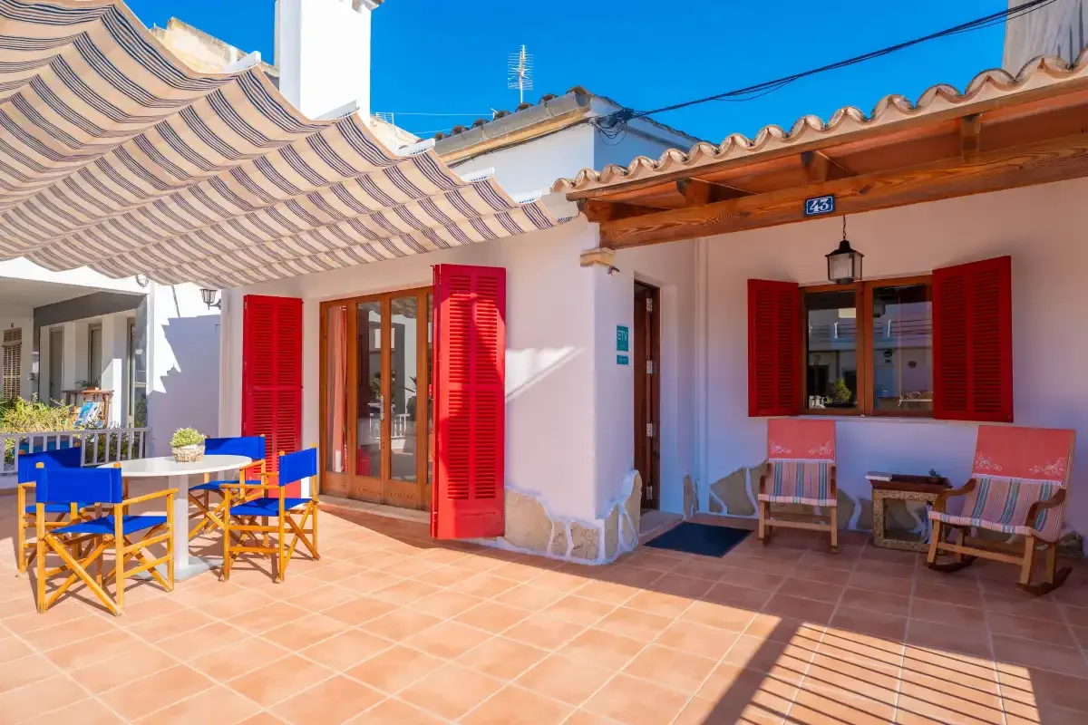 Un patio soleado con suelo de baldosas, contraventanas rojas, asientos y un toldo a rayas.
