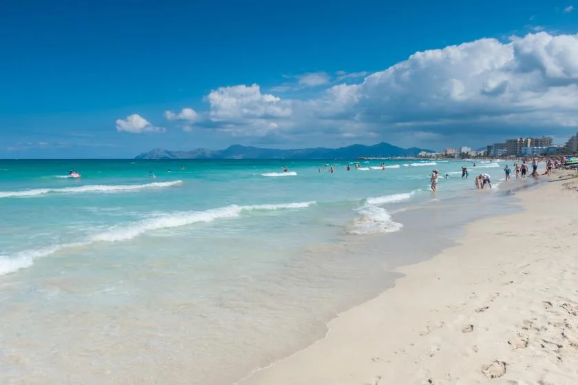 Una playa de arena bordea aguas turquesas, con montañas visibles al otro lado de la bahía.