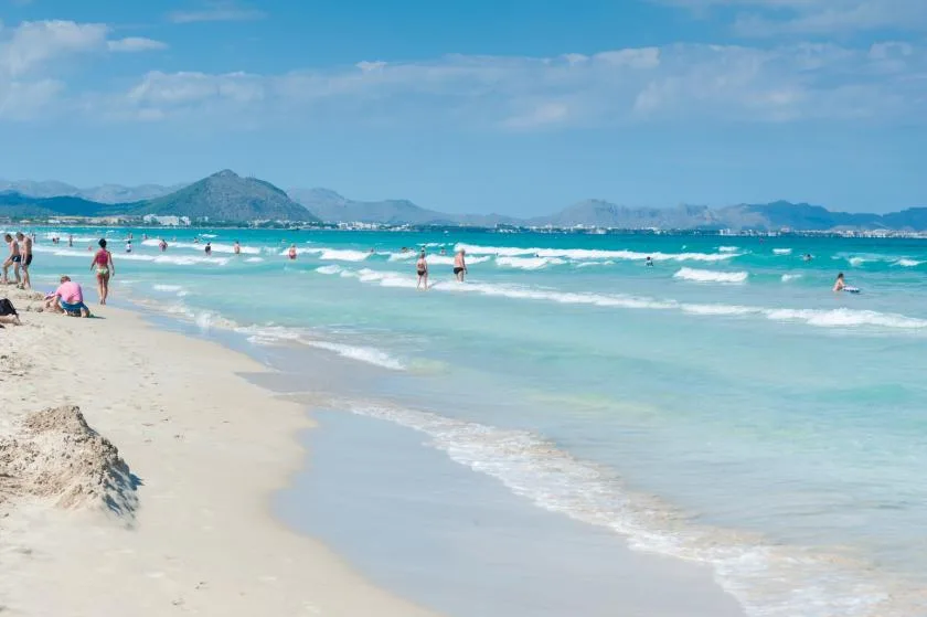 Una playa de arena se encuentra con aguas turquesas, con bañistas y montañas lejanas.