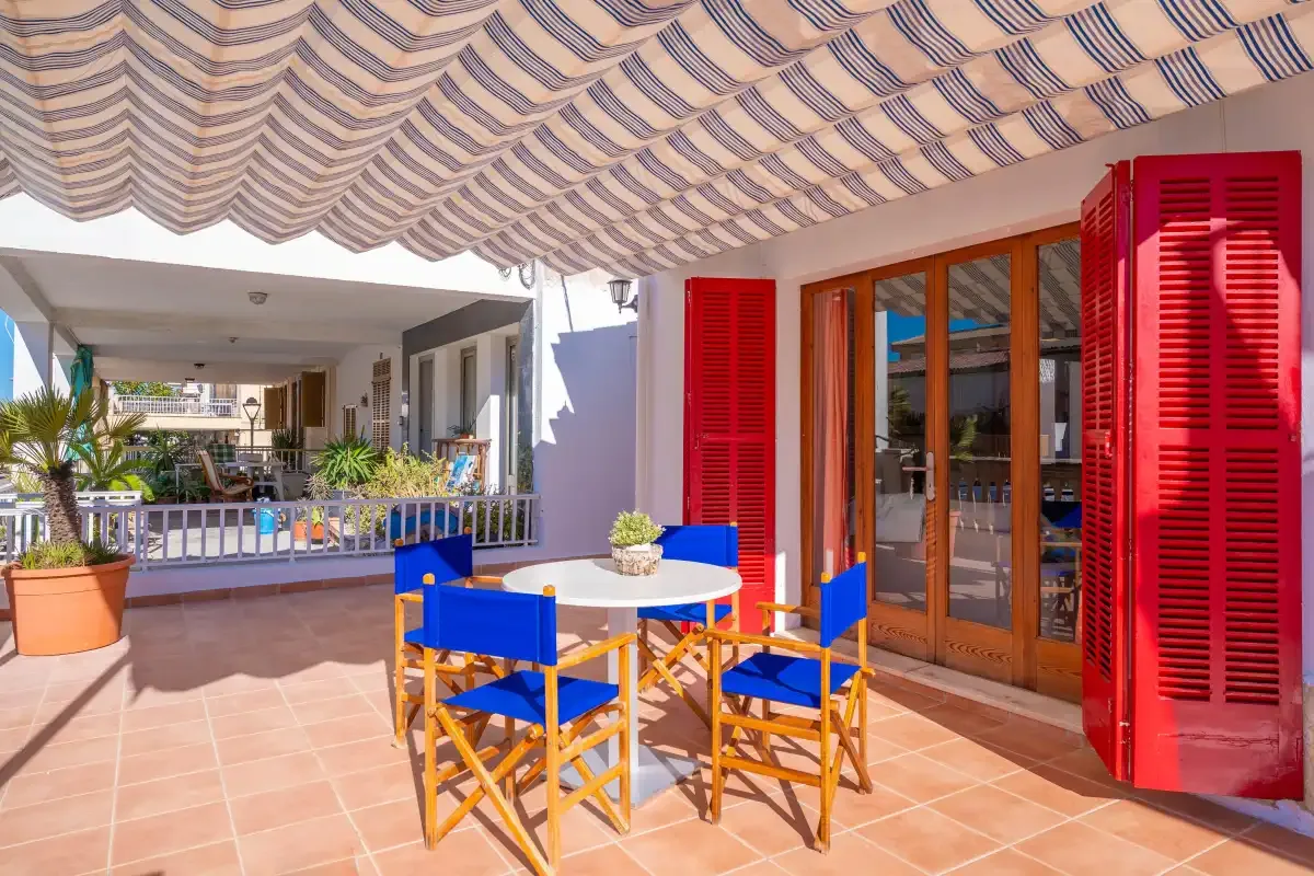 Una terraza sombreada con una mesa, asientos y contraventanas rojas abiertas.