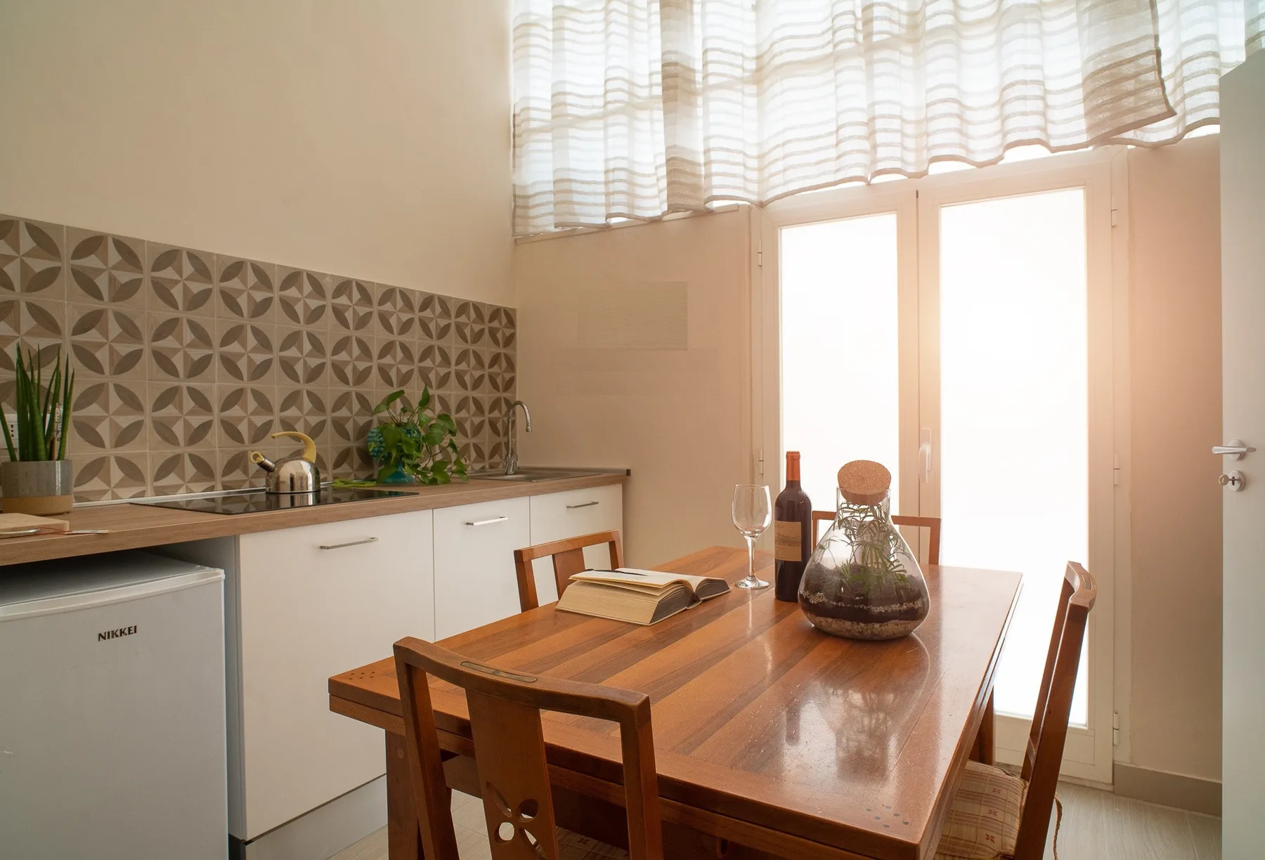 A dining area with a wooden table beside a small kitchen and bright windows.