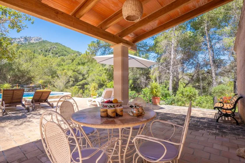 A covered outdoor dining area with a table and seating overlooks a pool and wooded hills.