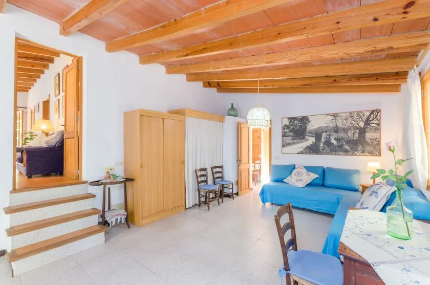 A bright living room has blue seating, a dining table, and exposed wooden ceiling beams.