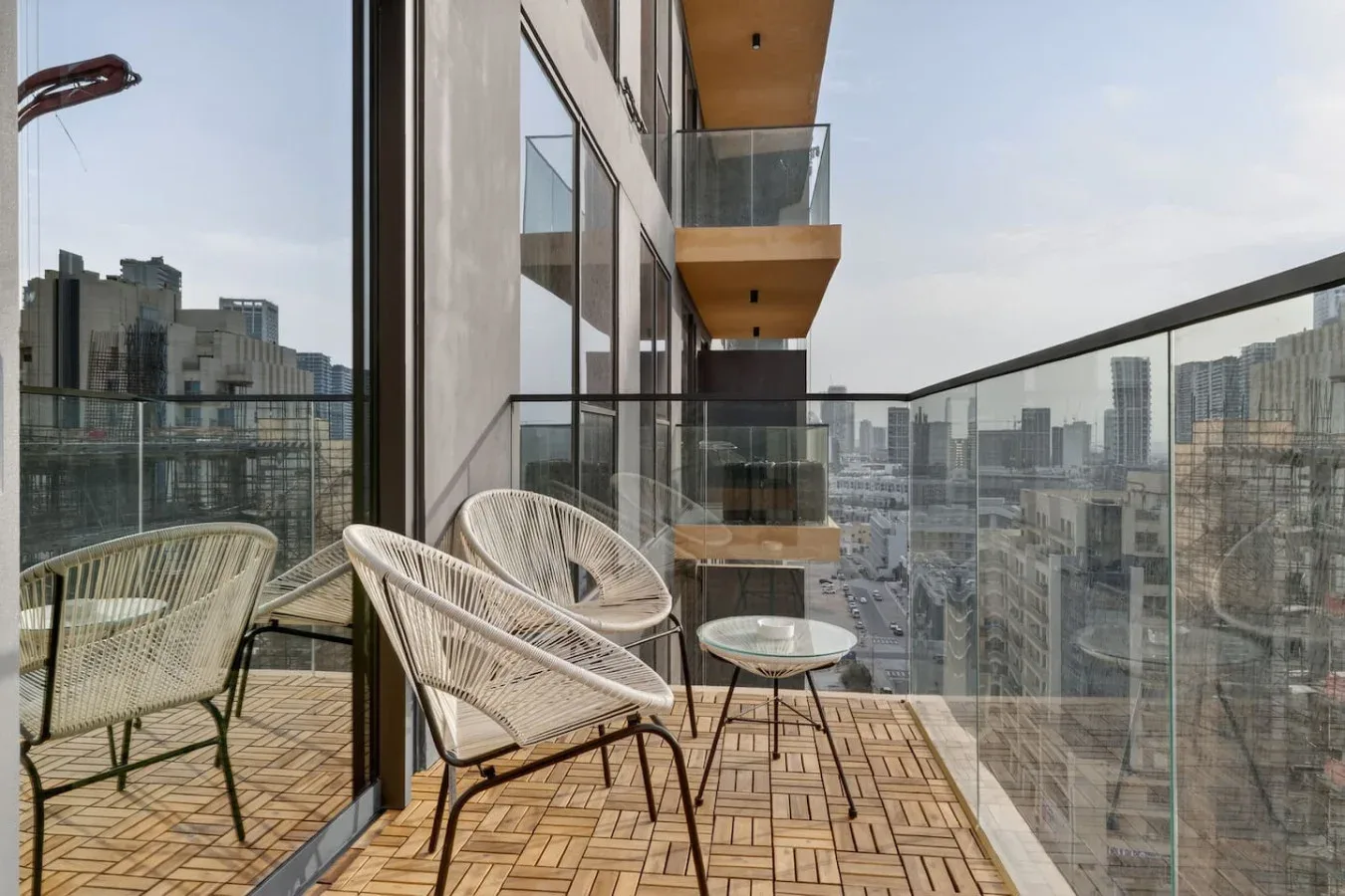 Una zona de estar en un balcón con asientos tejidos y una barandilla de vidrio con vistas a la ciudad.