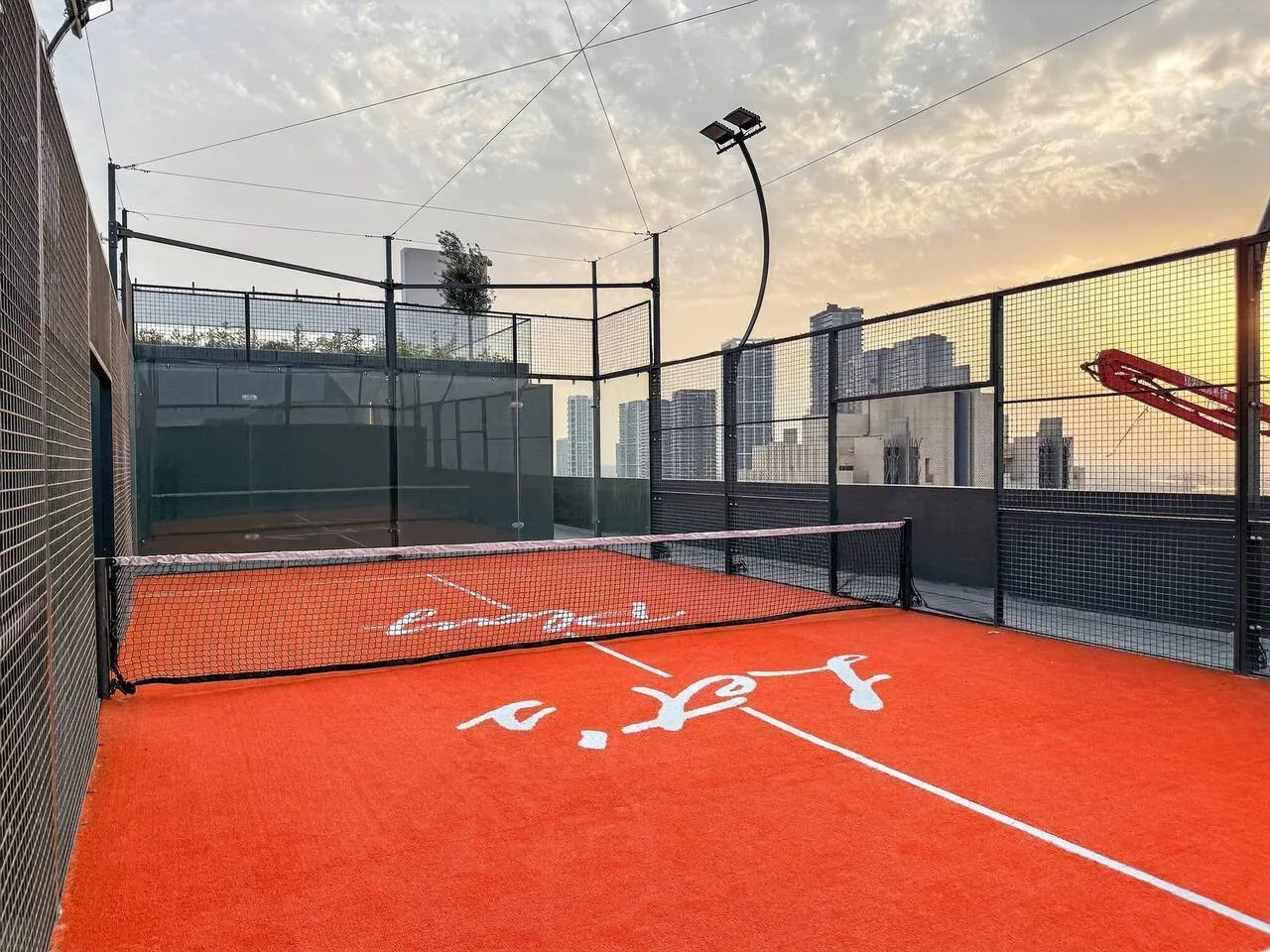 Una pista de pádel al aire libre con superficie naranja y edificios al fondo.