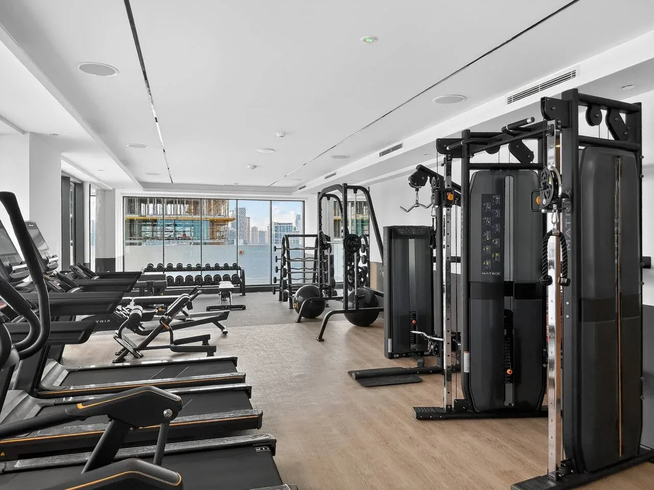 Un gimnasio con máquinas de ejercicio y una fila de cintas de correr junto a grandes ventanas.