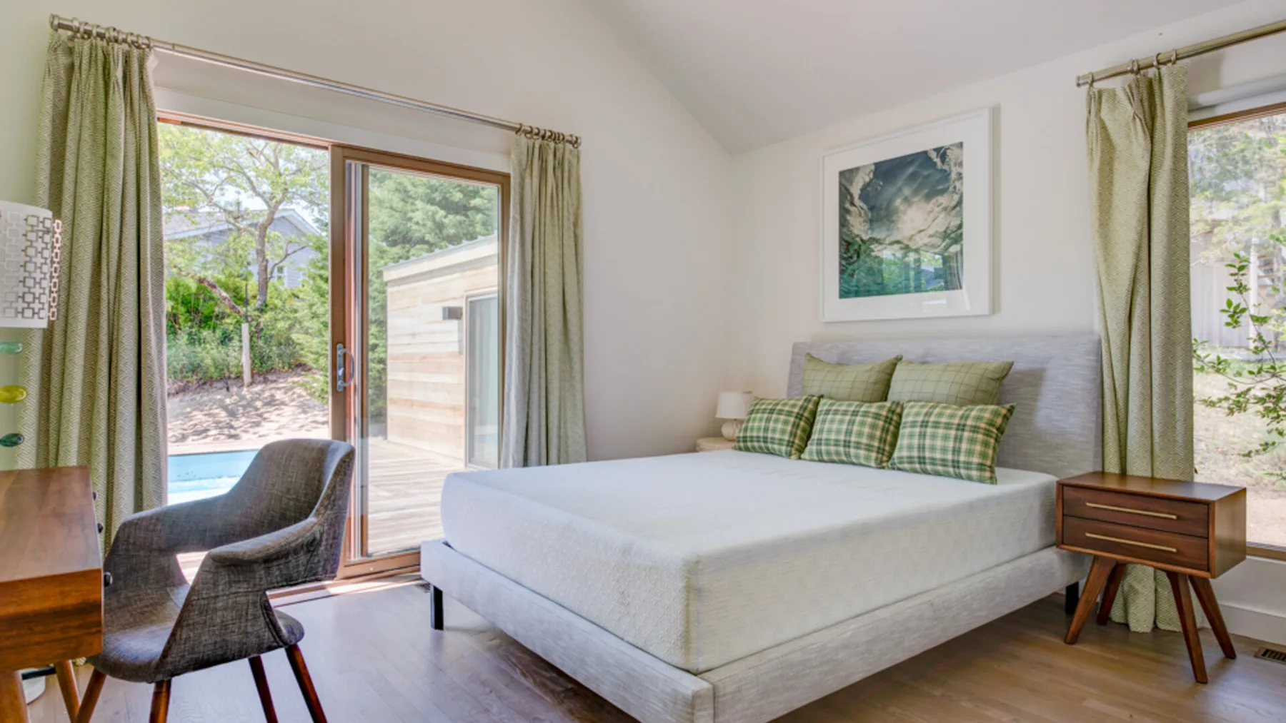 A bright bedroom with a bed, plaid pillows, and large windows overlooking greenery.