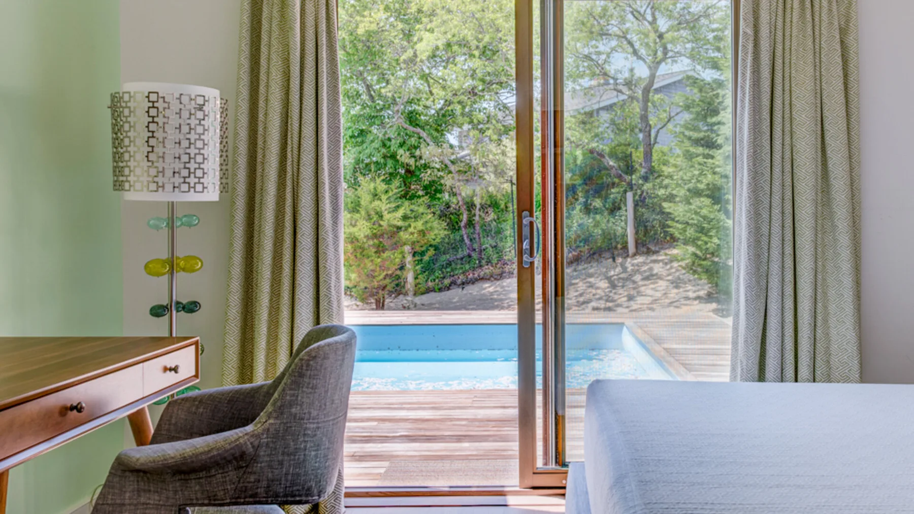 A bedroom opens onto a pool and wooden deck through large glass doors.