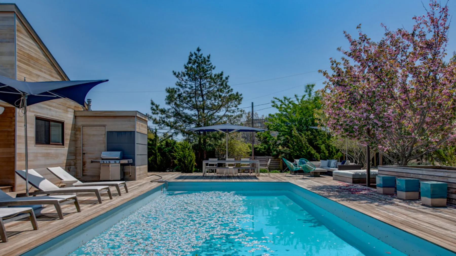 An outdoor swimming pool is surrounded by wooden decking, loungers, and trees.