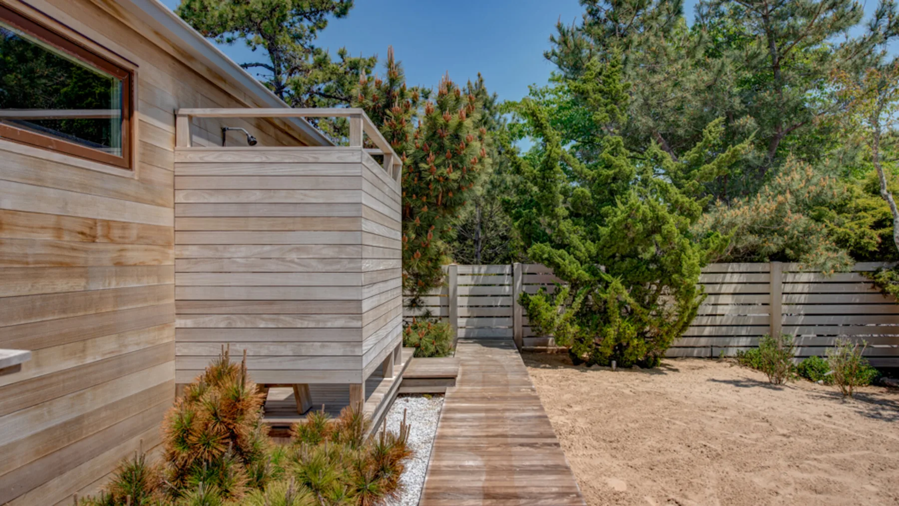 A wooden deck runs beside a wood-clad building, with trees and a sandy yard.