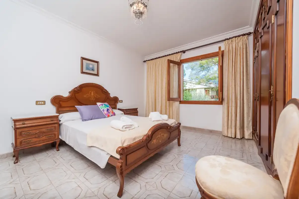 A bedroom with a carved wooden bed, bedside table, and curtained window.