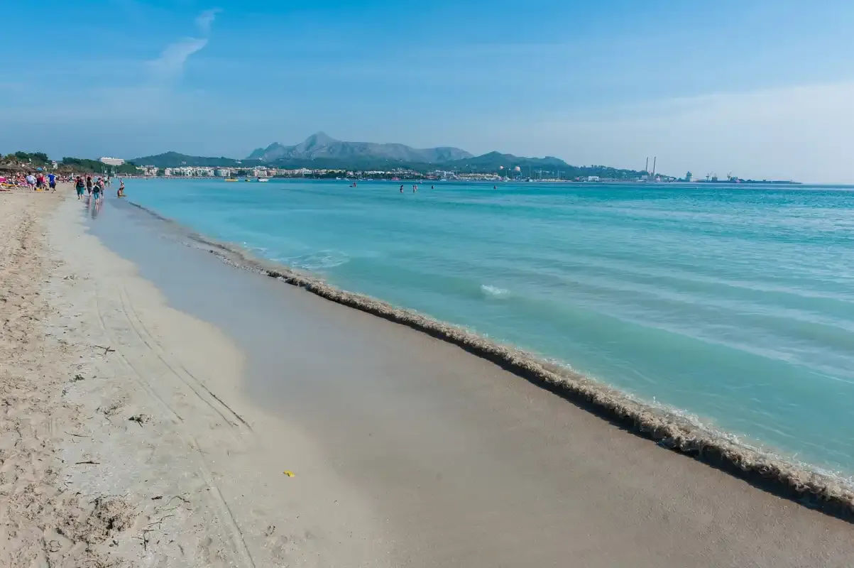 A sandy beach borders clear blue water, with distant mountains along the coast.