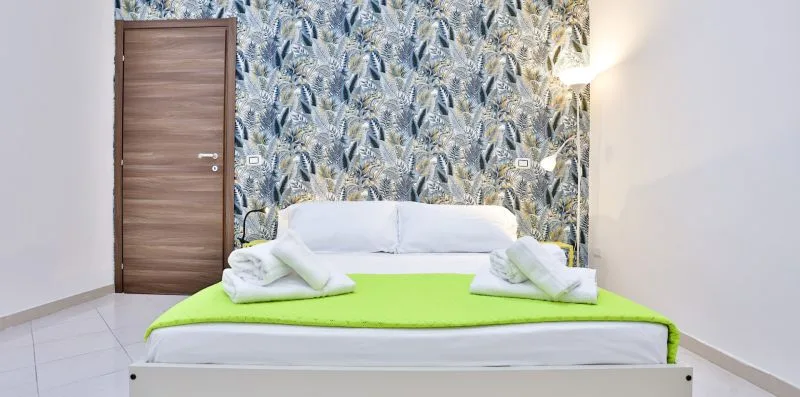 A bedroom with a bed covered by a bright green blanket and botanical wallpaper.
