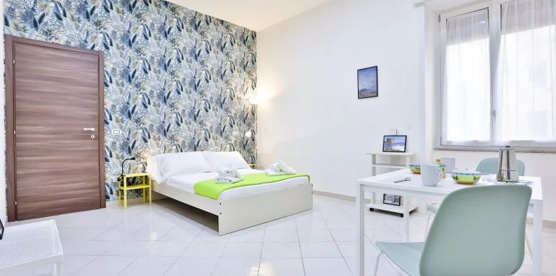 A bedroom with a bed beside a desk and large windows.