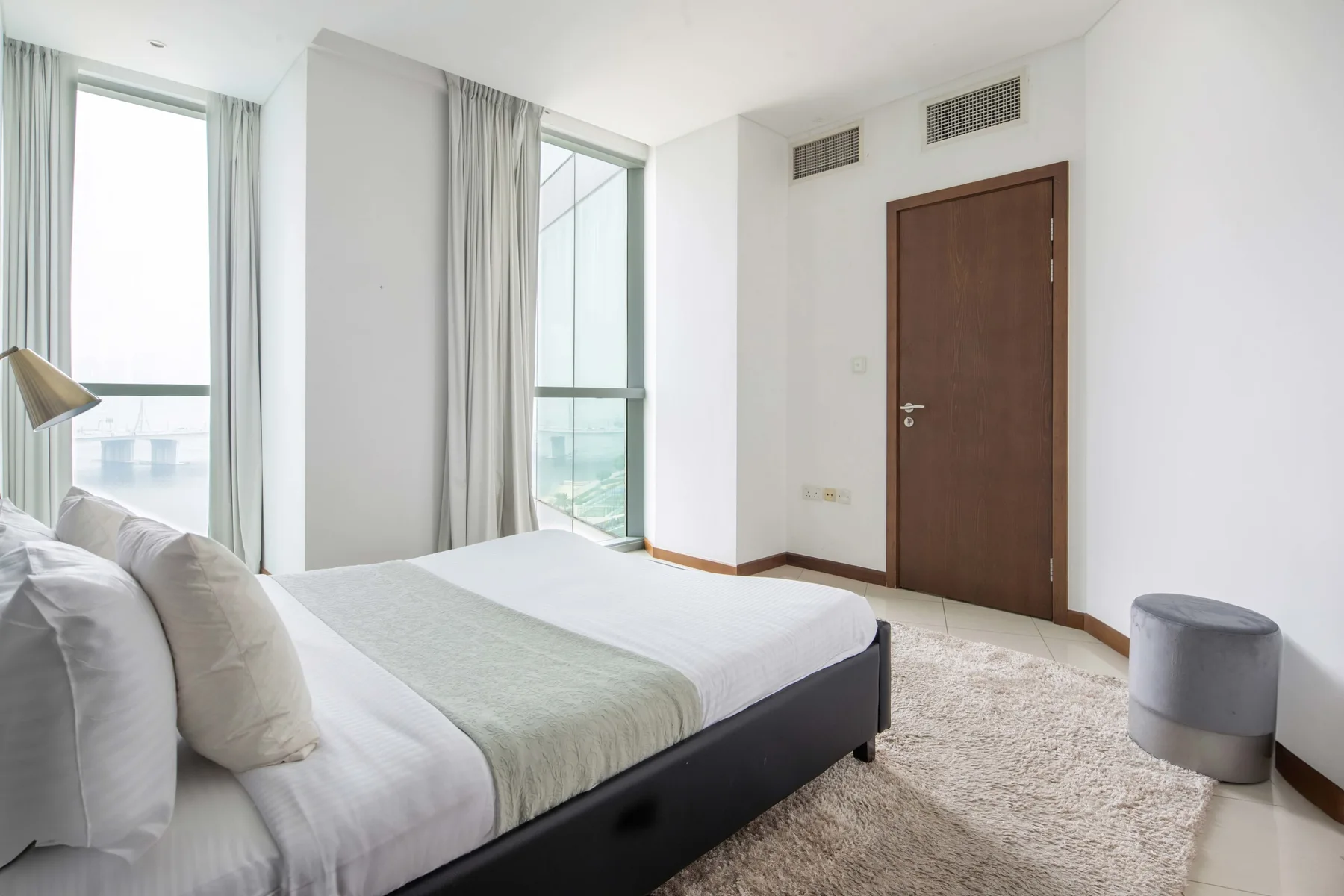 Un dormitorio luminoso con una cama junto a ventanas altas y una puerta marrón.