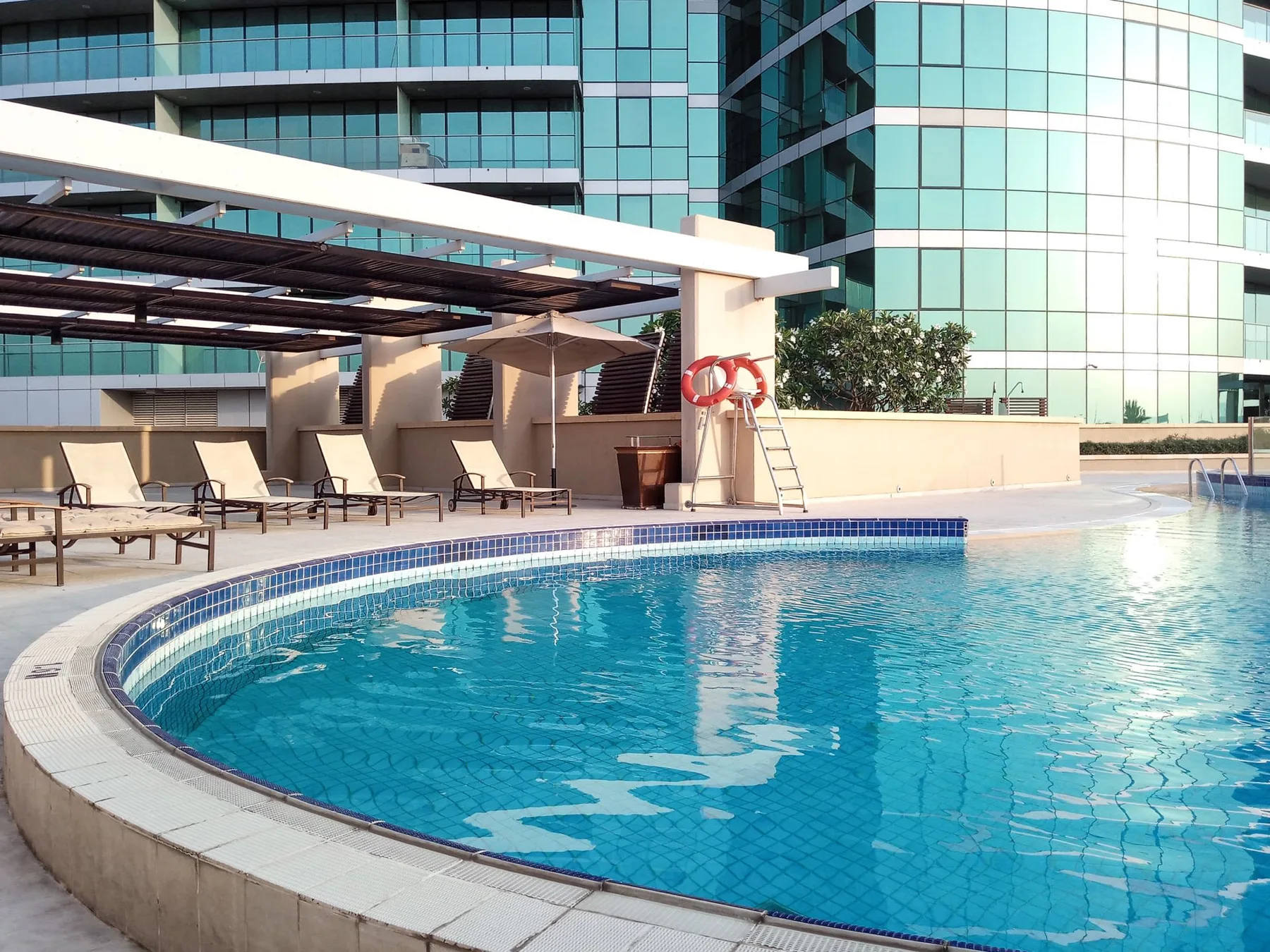 Una piscina exterior curva está junto a un edificio acristalado y tumbonas a la sombra.