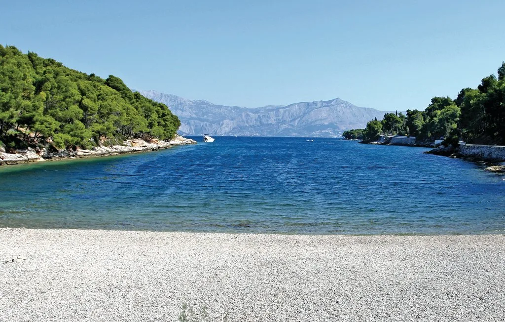 A pebbly beach borders a blue inlet between green, tree-covered shores.