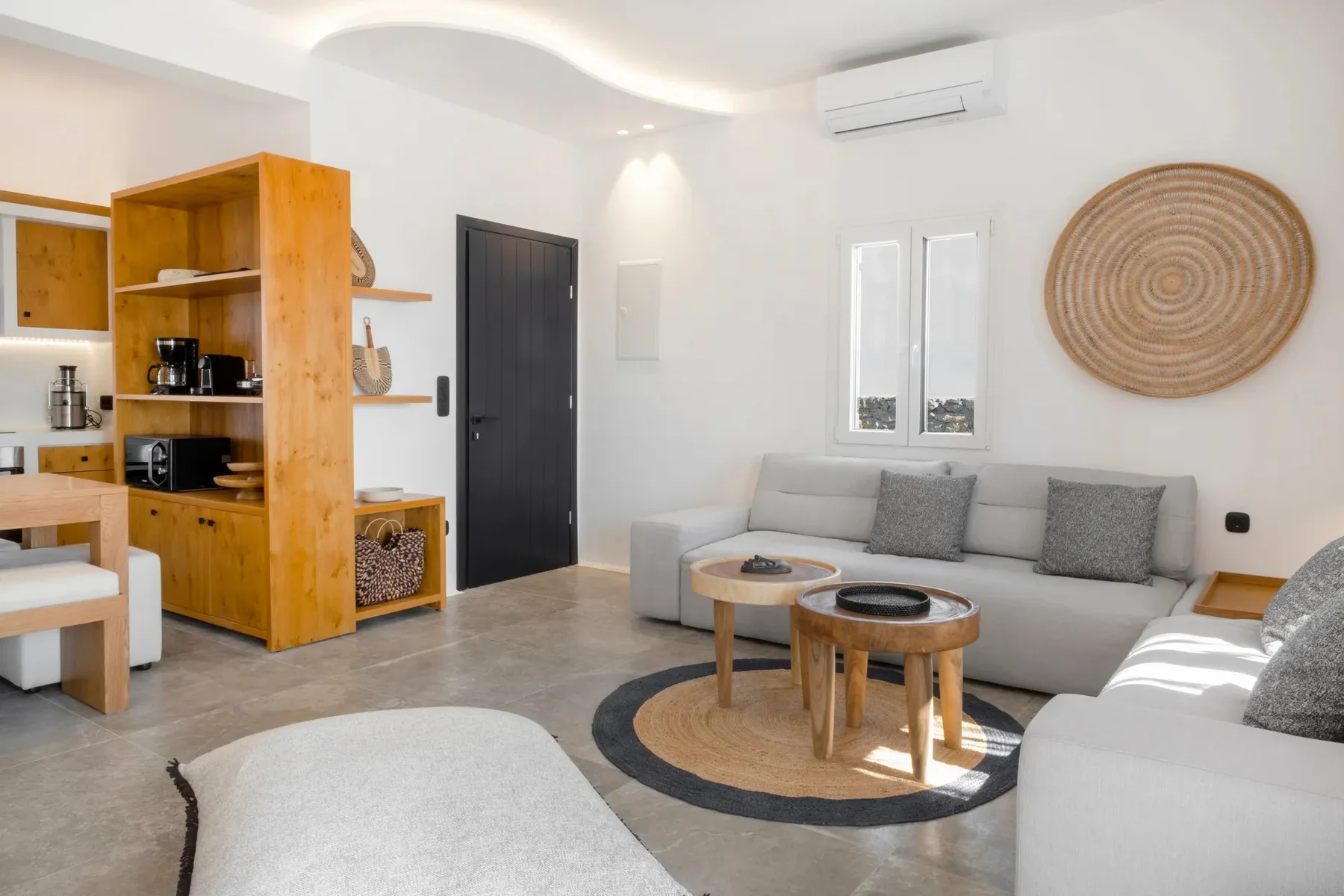 A bright living room with a gray sofa, wooden tables, and a compact kitchen.