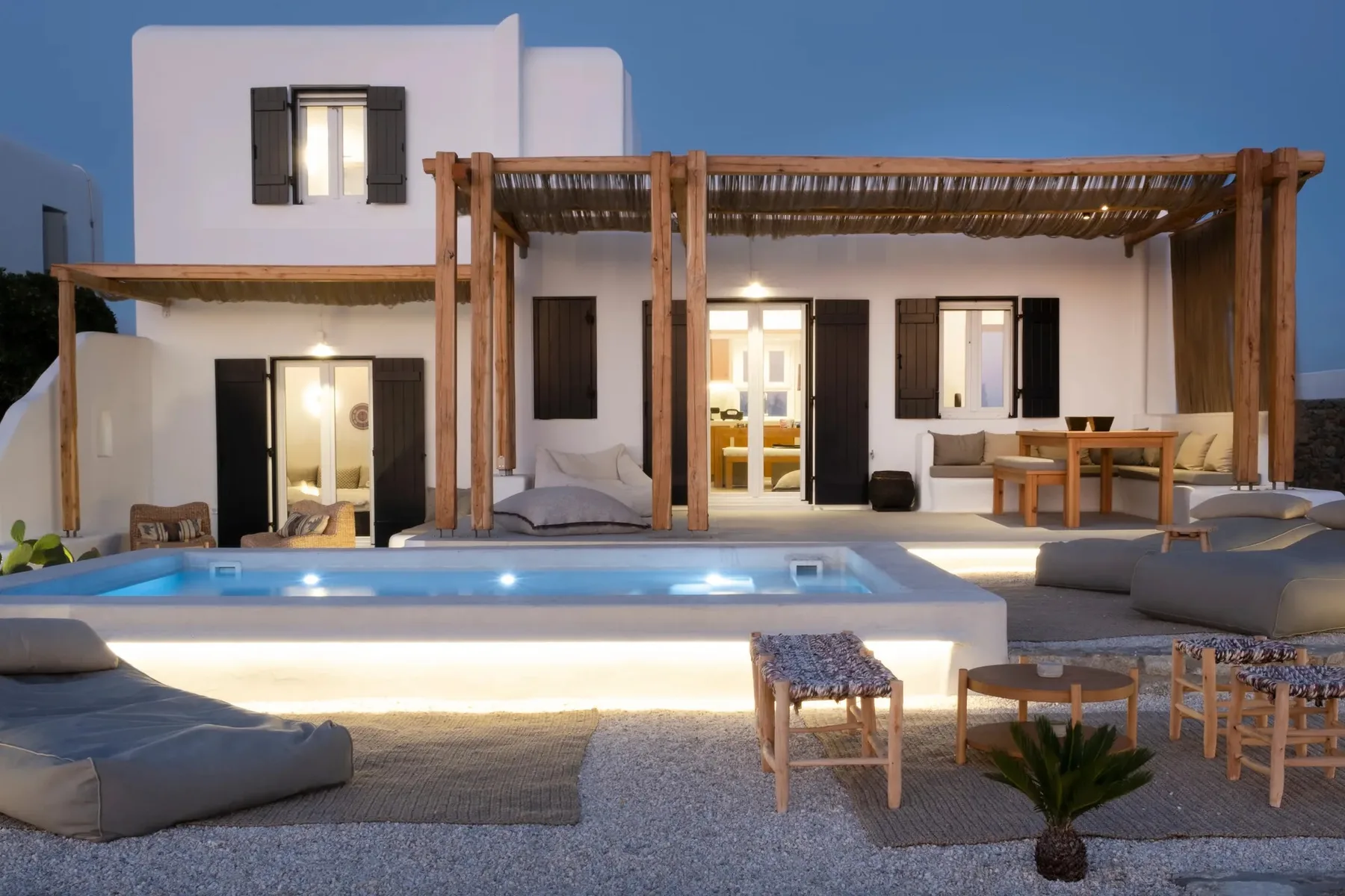 A white villa opens onto a lit swimming pool and outdoor seating.