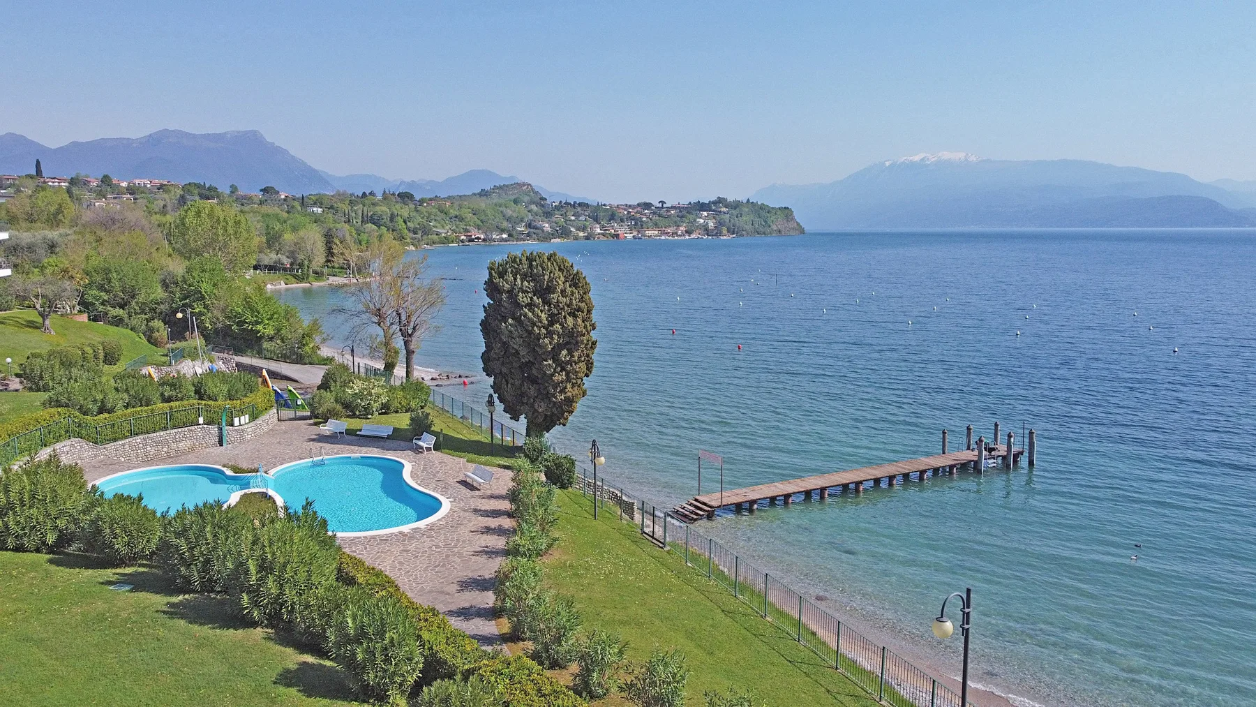 A lakeside garden with a swimming pool and a wooden pier extending into the water.