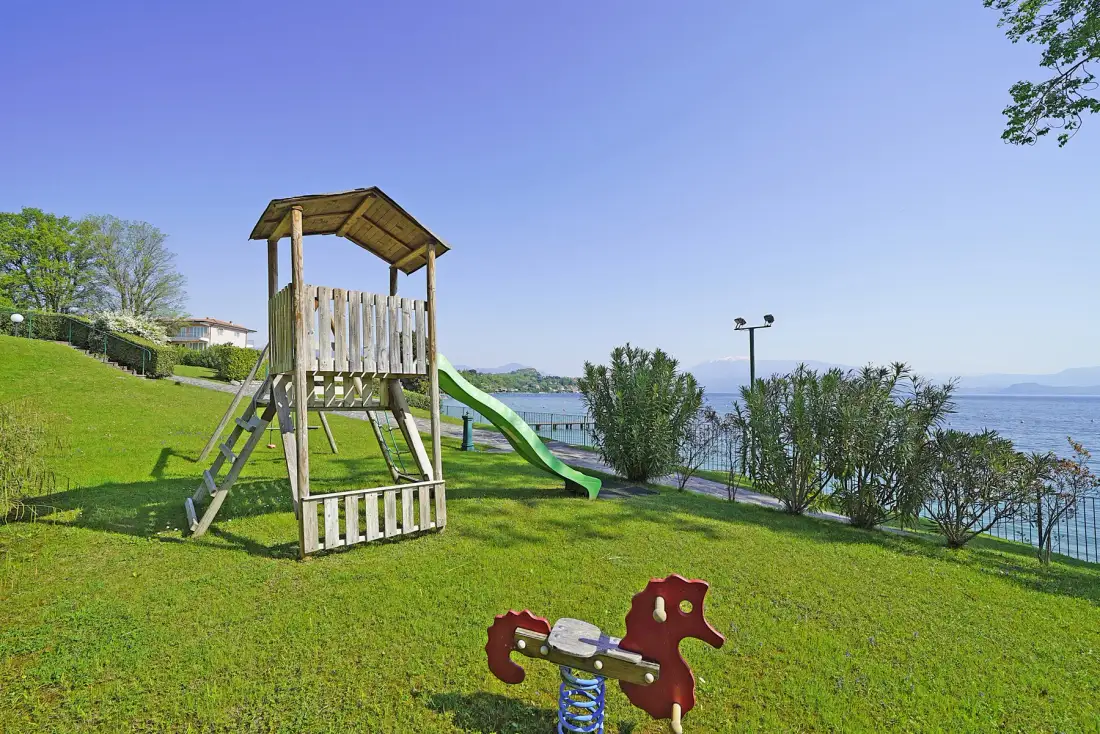 A grassy outdoor playground with a wooden play structure and slide beside the water.