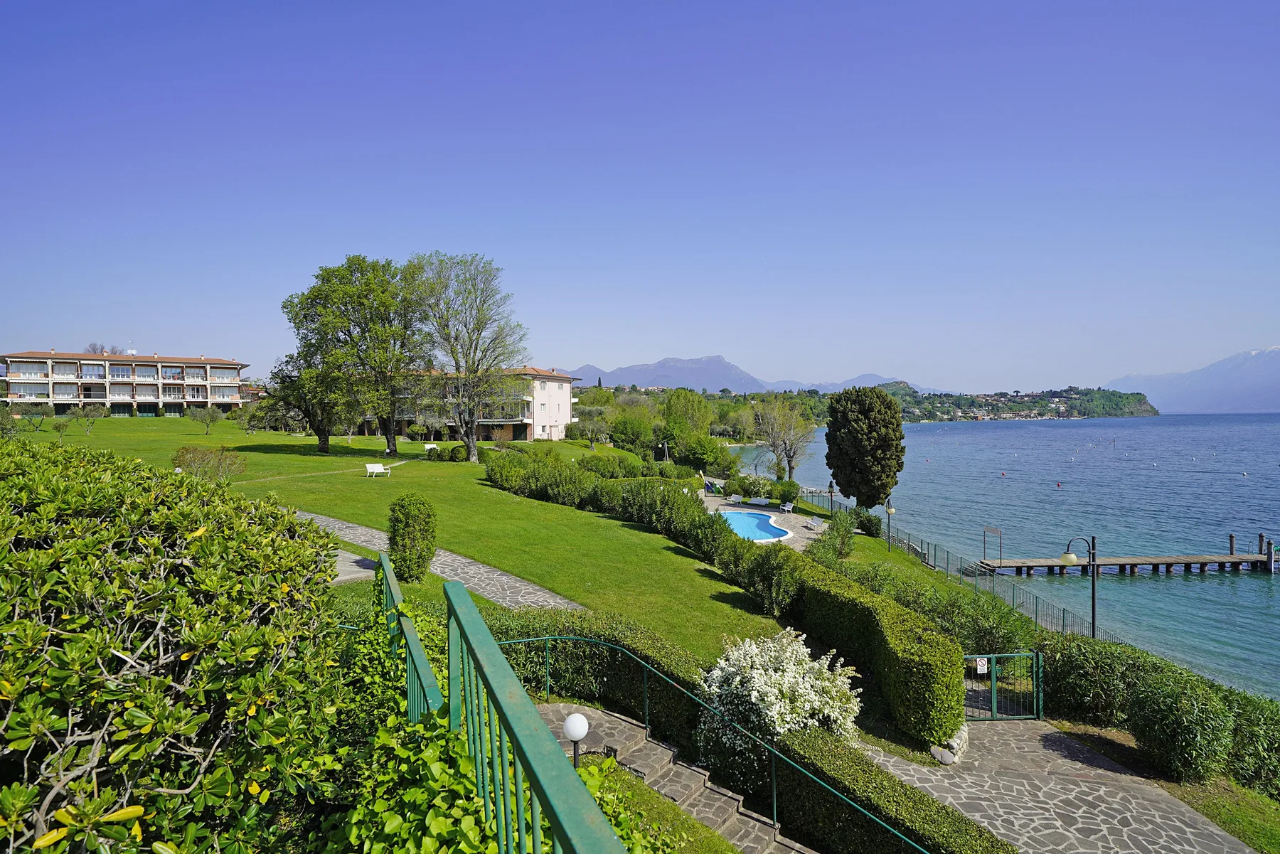 A landscaped waterfront grounds overlook a lake, with a paved path and distant mountains.
