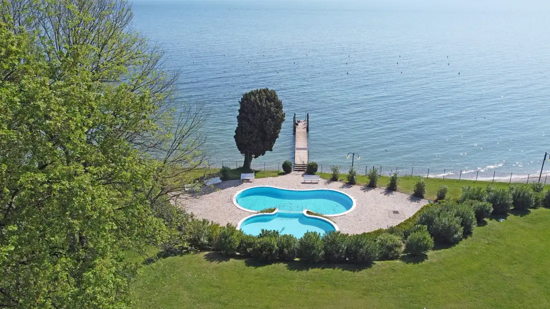 A kidney-shaped pool sits in a landscaped area beside the water.