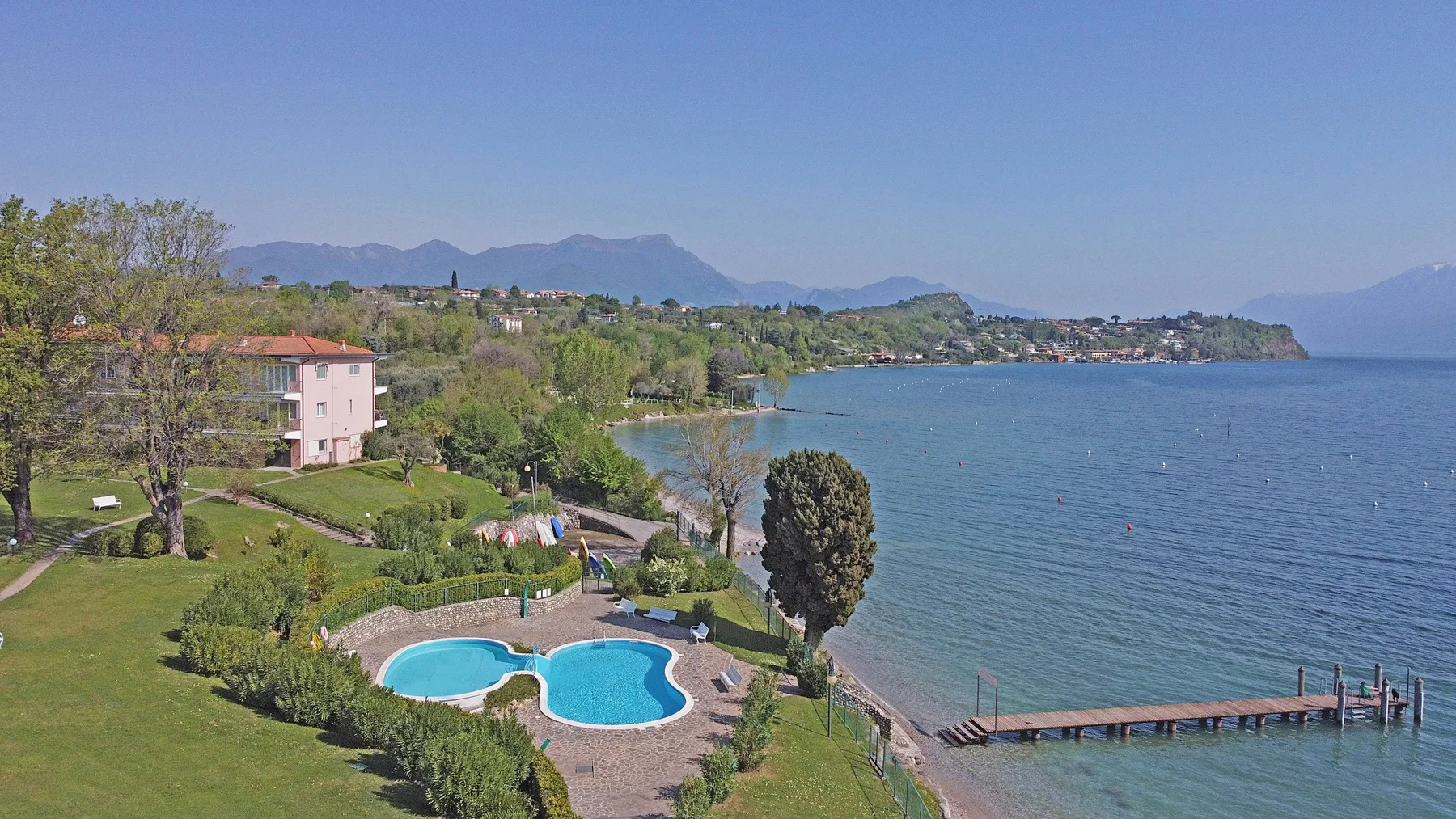 A landscaped waterfront with a swimming pool, a pier, and mountains across the lake.