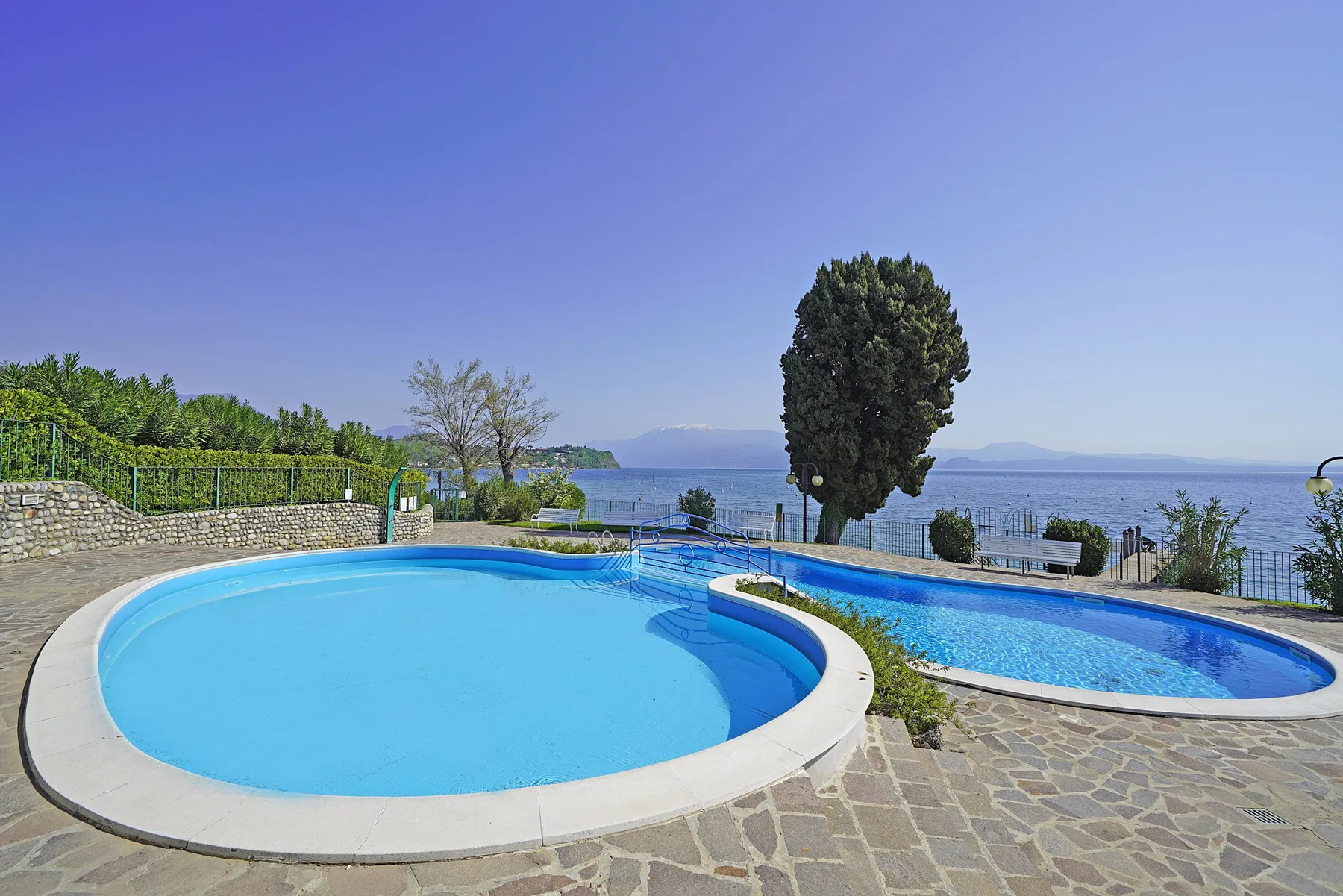 A swimming pool beside a second pool, with trees and a lake in the background.