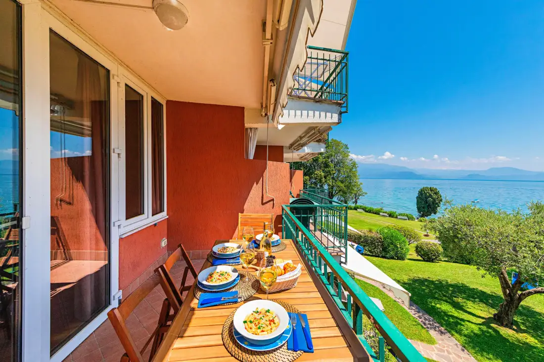A balcony dining area overlooks a green garden and a lake.
