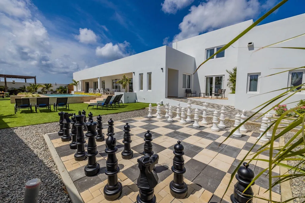 Modern villa featuring an oversized chess set on the lawn, where sun-drenched days invite strategic play beside the pool amid palms and endless blue skies.