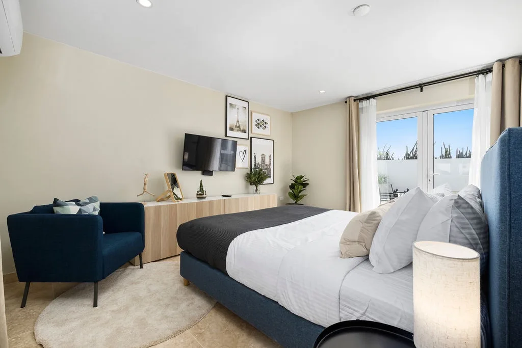 Sink into crisp linens and plush pillows in this serene bedroom. Natural light streams through balcony doors, while modern touches and a cozy seating nook invite you to unwind.