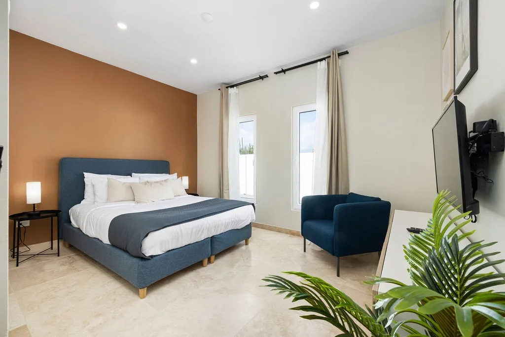 Sink into the plush king bed beneath the warm accent wall, relax in your cozy armchair, and enjoy modern comfort in this thoughtfully designed bedroom where every detail invites you to unwind.