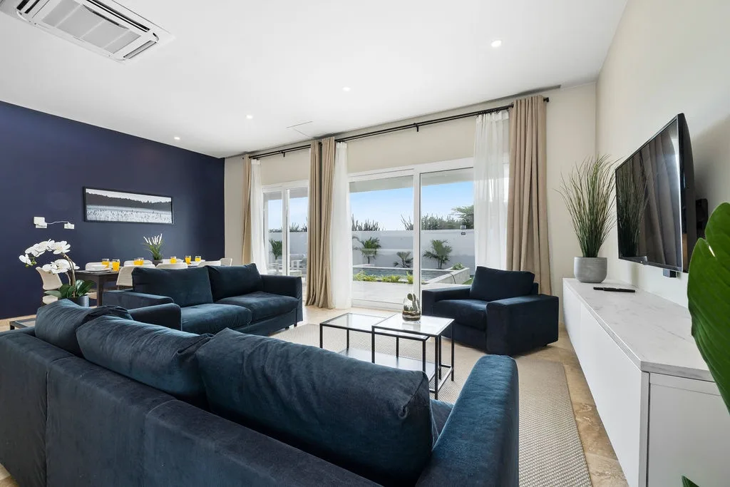 Sink into plush navy seating as sunlight streams through floor-to-ceiling windows, framing serene garden views. Your open-plan living and dining space invites relaxation with modern comfort and coastal style.