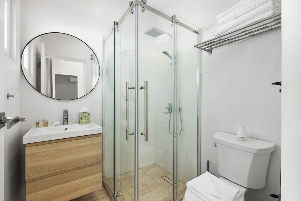 Your modern ensuite bathroom features a spacious walk-in shower with rainfall showerhead, sleek vanity with round mirror, and fresh towels waiting for your arrival.