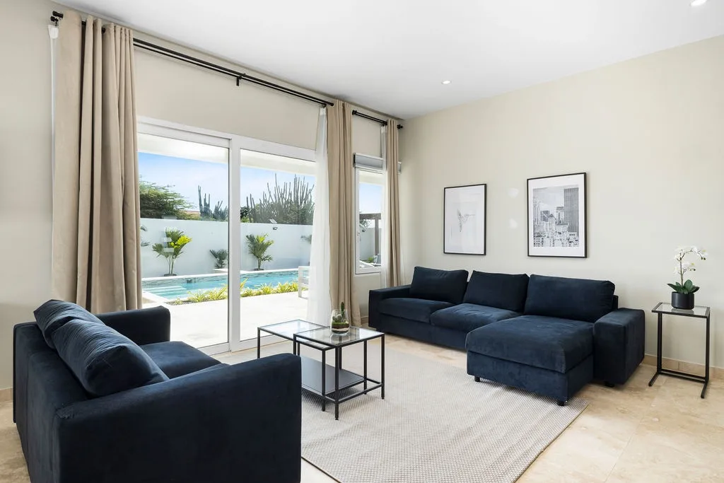 Sink into plush navy sofas in your sunlit living room, where floor-to-ceiling glass doors frame sparkling pool views and a serene outdoor oasis. Perfect for unwinding after exploring the island.