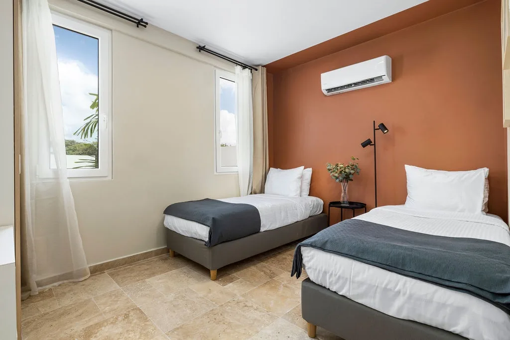 Your comfortable twin bedroom features crisp linens, contemporary styling, and air conditioning to keep you cool. Natural light streams through the windows, highlighting the warm terracotta accent wall.