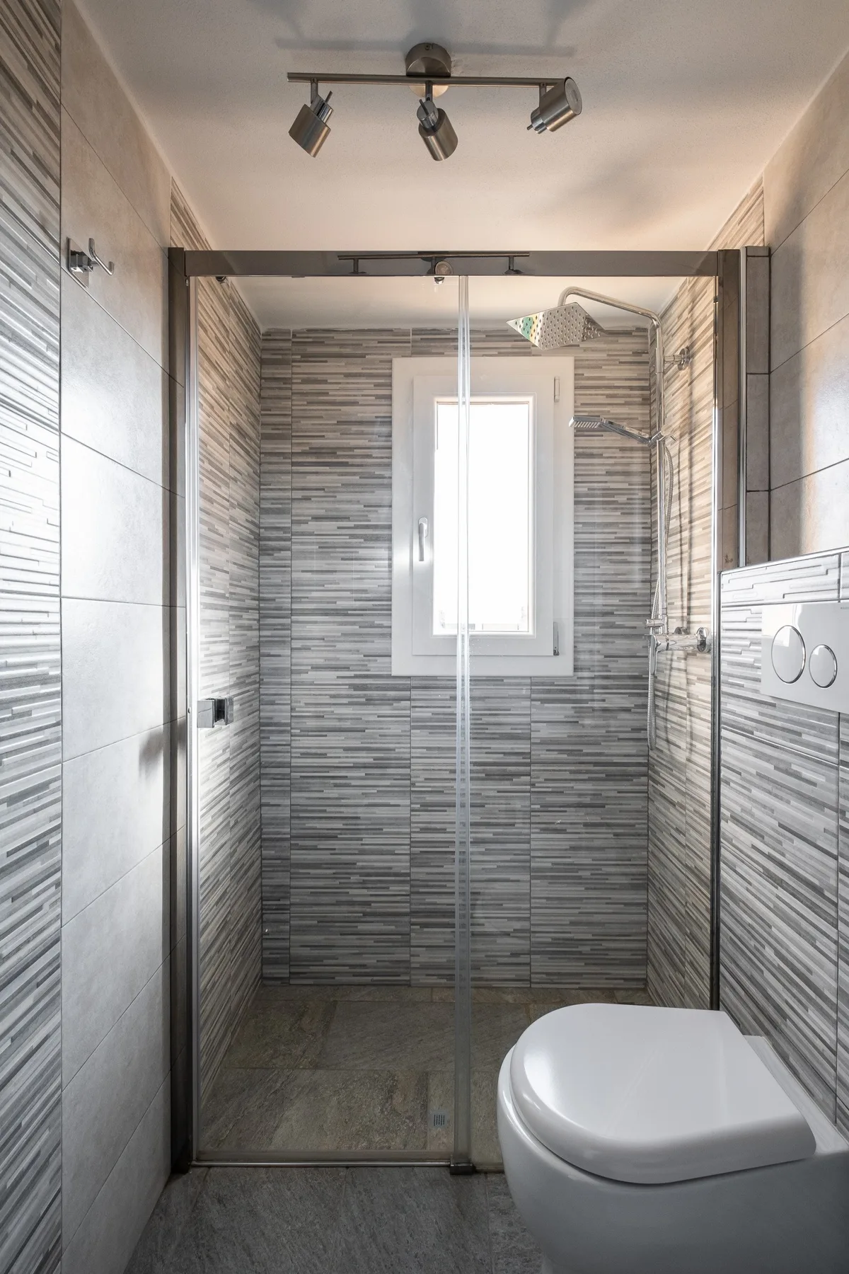 A tiled bathroom with a glass shower and a toilet beside a window.