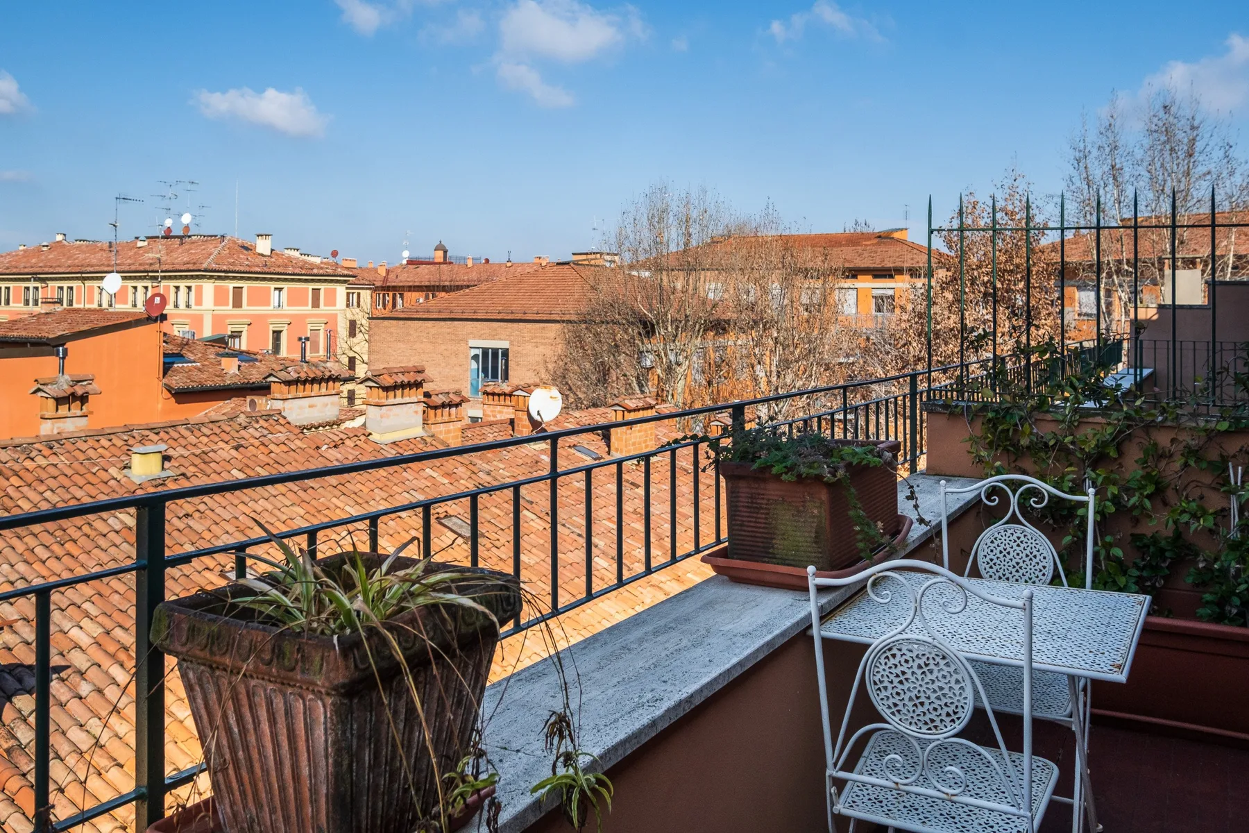 Una terraza en la azotea da a edificios cercanos, con asientos metálicos y plantas en macetas.