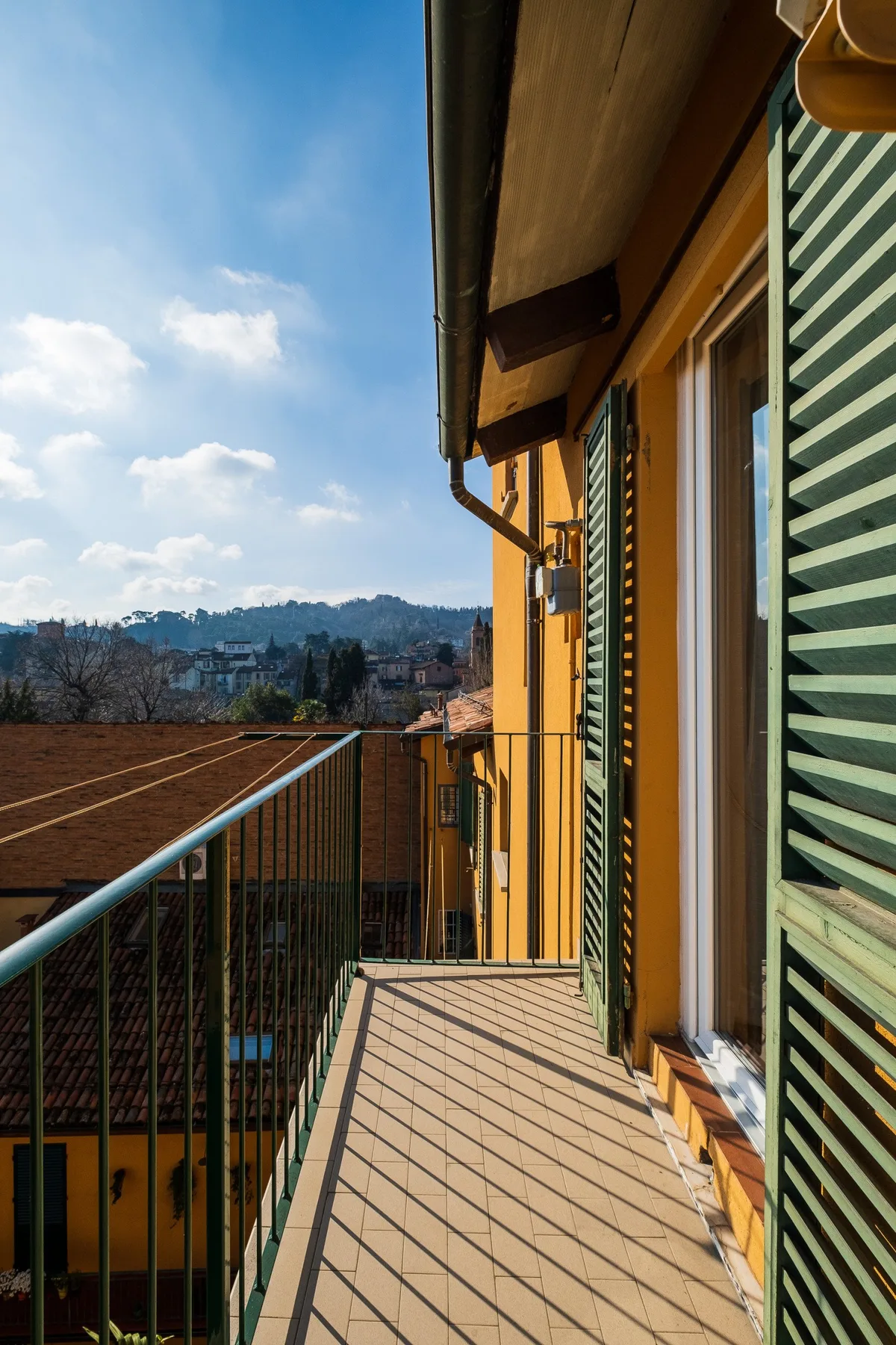 A balcony beside a yellow building overlooks rooftops and distant hills.