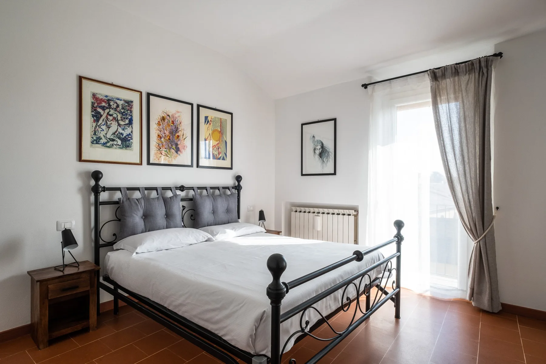 A bedroom with a metal bed, framed artwork, and a bright curtained window.