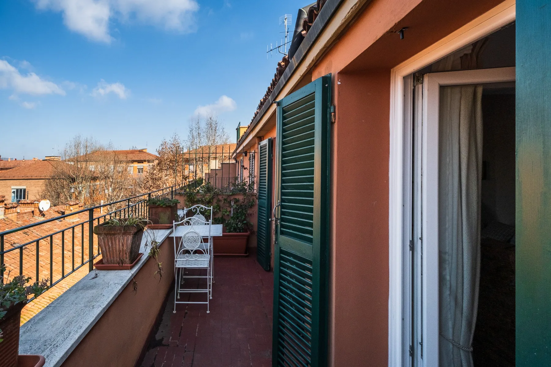 A narrow balcony runs alongside a building, with green shutters and potted plants.