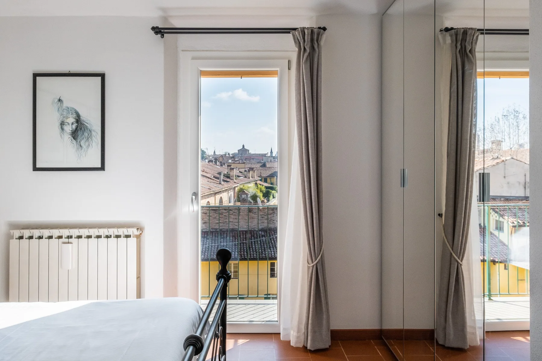 A bedroom with a bed, radiator, and a window overlooking nearby rooftops.