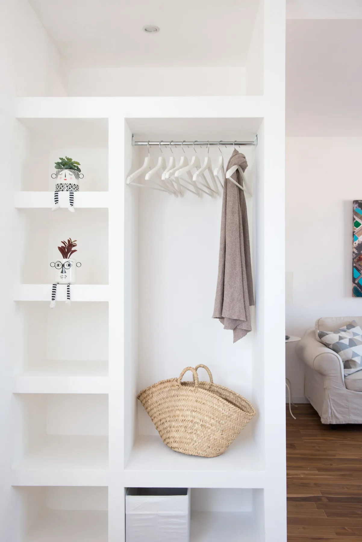 A white open shelving unit holds hanging clothes, small plants, and a woven basket.