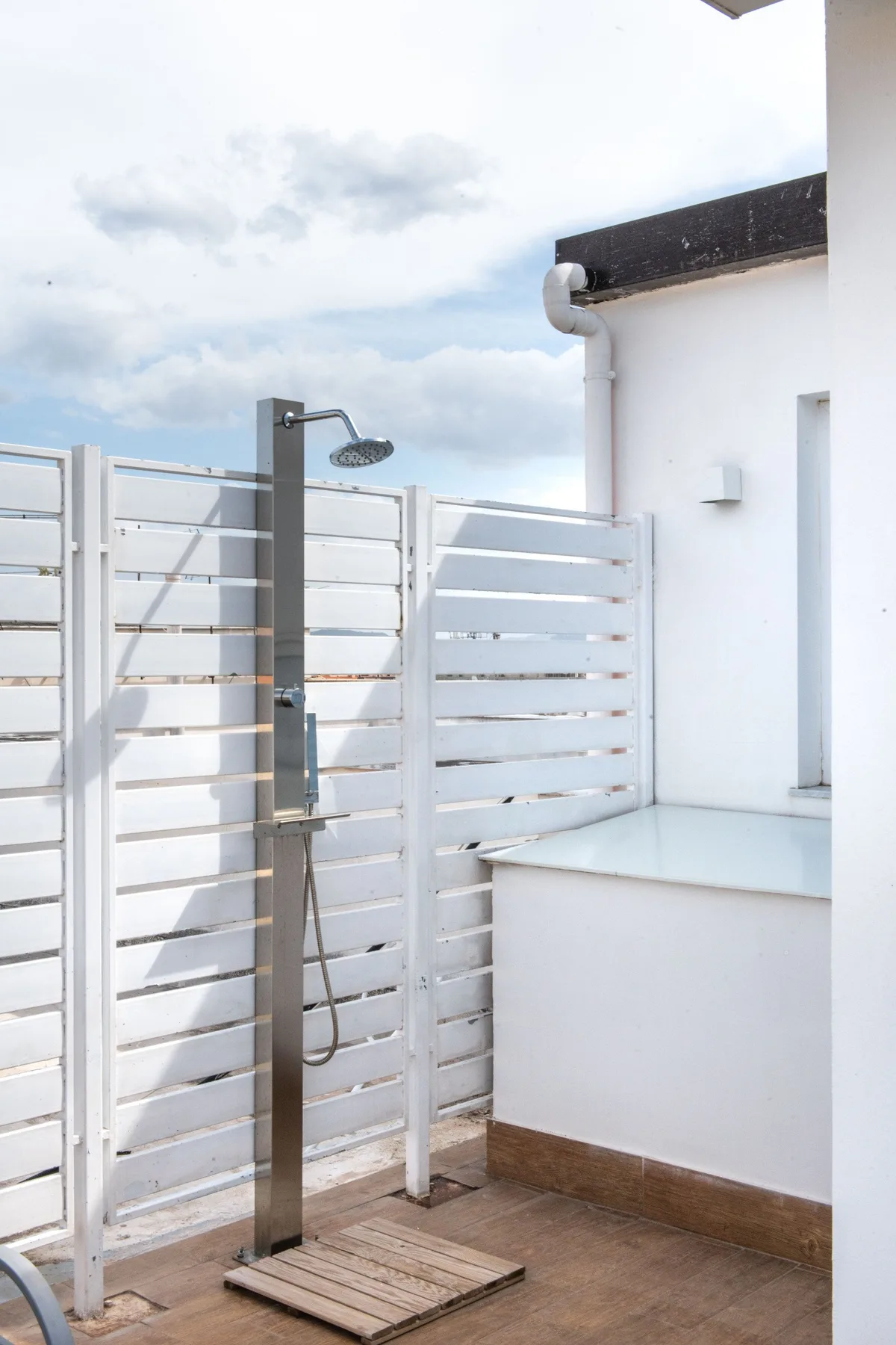 An outdoor shower stands beside a white wall and horizontal privacy panels.