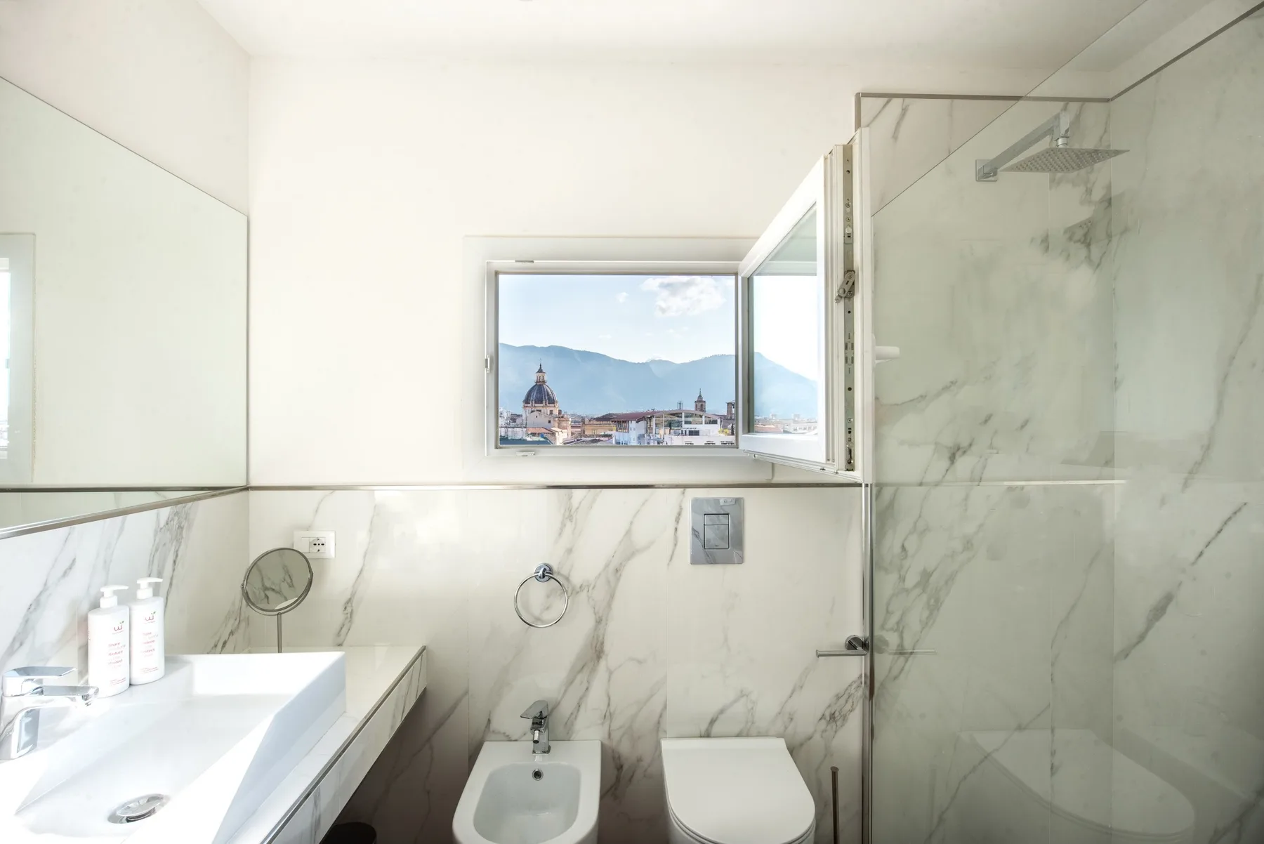 A bright bathroom with a sink, toilet, and an open window overlooking the city.