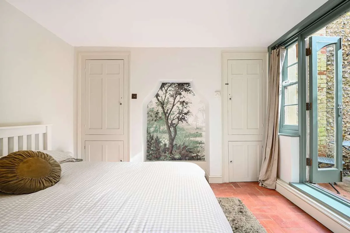 Un dormitorio con una cama junto a ventanas altas y un panel pintado en la pared.