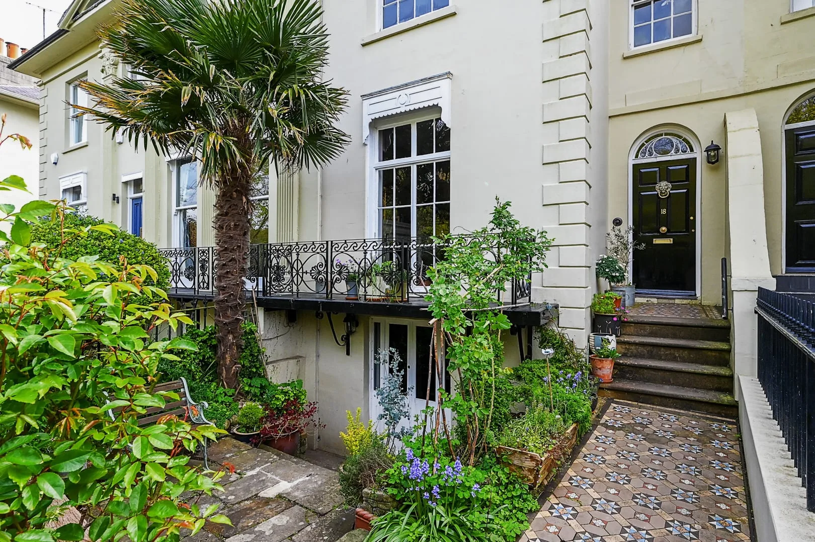 A leafy courtyard garden beside a pale building, with a balcony and paved walkway.