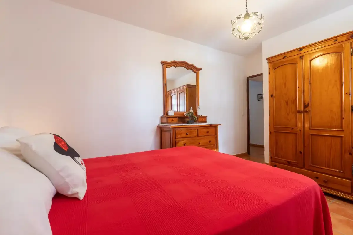 A bedroom with a red-covered bed, wooden wardrobe, and dresser.