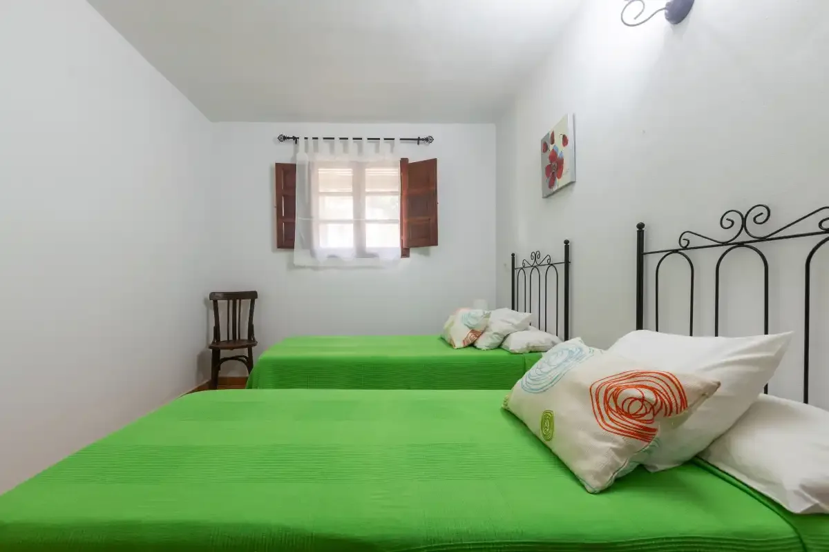 A bright bedroom has green bedding, iron bed frames, and a window with open shutters.